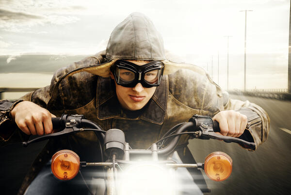 Bild vergrößern: Das Bild zeigt einen jungen Mann mit einer Fliegerbrille und einer Pilotenfliegermtze in Leder der sich weit auf den Lenker seines fahrenden Motorrads lehnt.